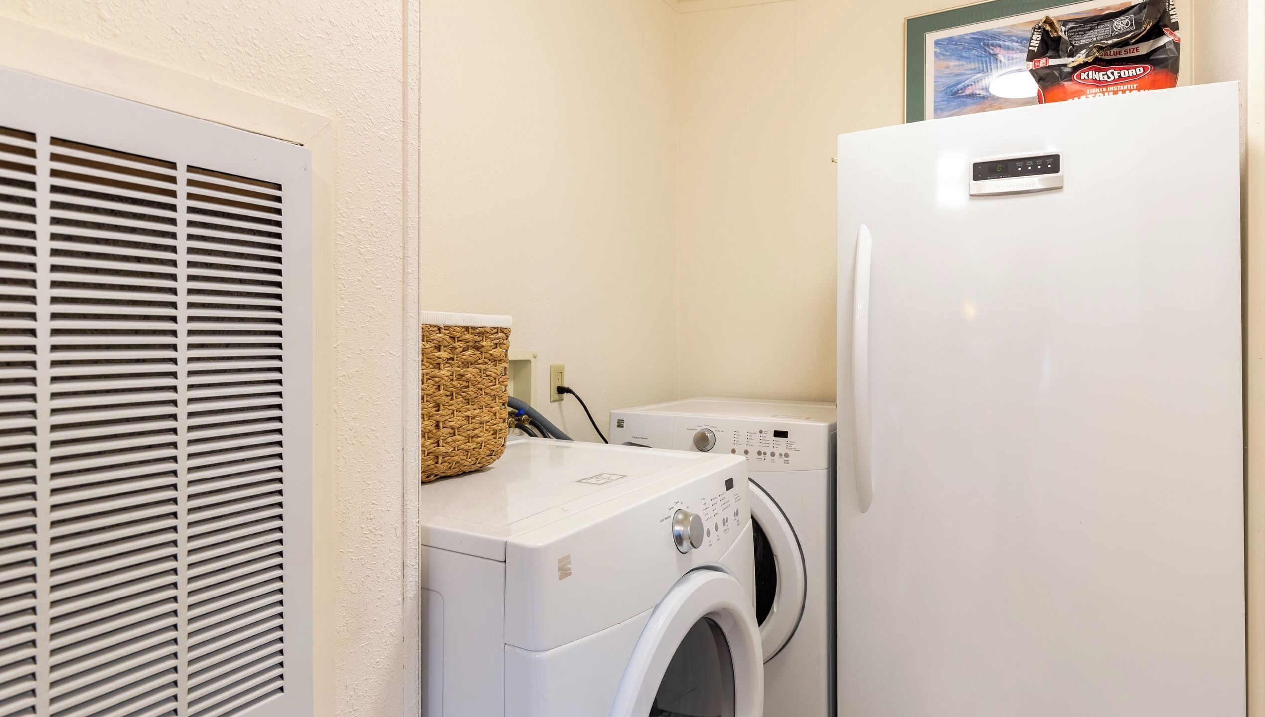 A laundry area with a white wall, a vent, a washing machine, a dryer, and a bag of Kingsford charcoal on top of the dryer.