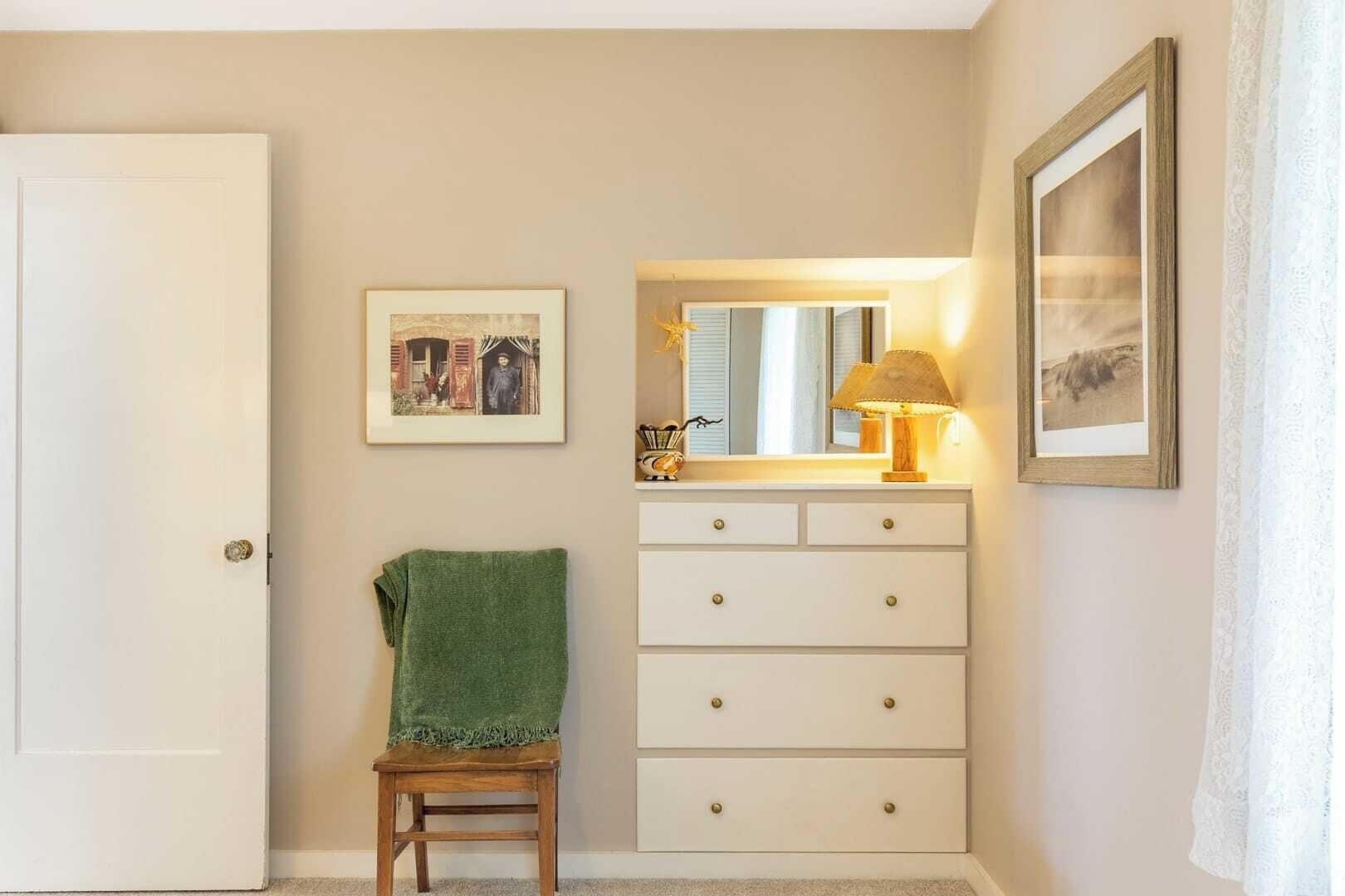 A room with a white door, a green towel on a wooden stool, a white dresser with drawers, a mirror, and two framed pictures on the wall.