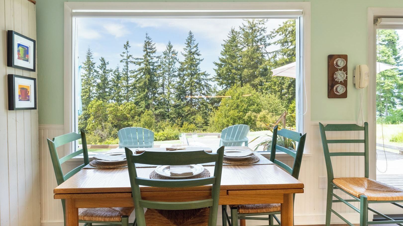Una mesa de comedor con sillas, una ventana con vista a árboles, y un teléfono en la pared.