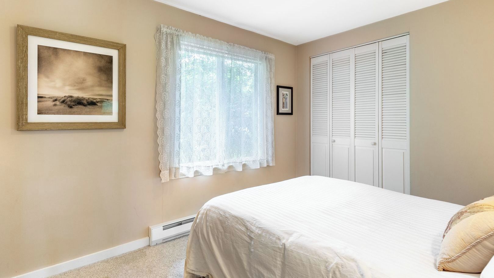 A bedroom with a bed, a window with lace curtains, a framed picture on the wall, and a white closet.