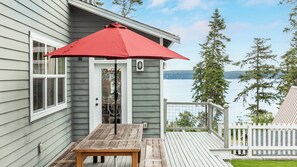 A wooden deck with a table and a red umbrella, overlooking a lake with trees and a house in the background. - The Lookout at Crane's Landing by Avantstay (Greenbank)