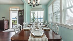 Une salle à manger avec une longue table dressée pour six personnes, un lustre et une vue par la fenêtre.
