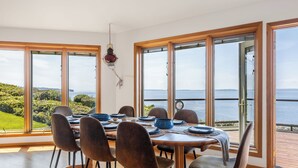 A dining area with a table set for six, large windows with a view of the sea, and a hanging light fixture. - The Majestic Blue View by Avantstay Waterfront (Freeland)