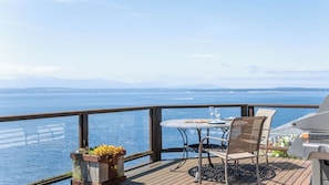 A deck with a table and chairs, overlooking a body of water and distant mountains.