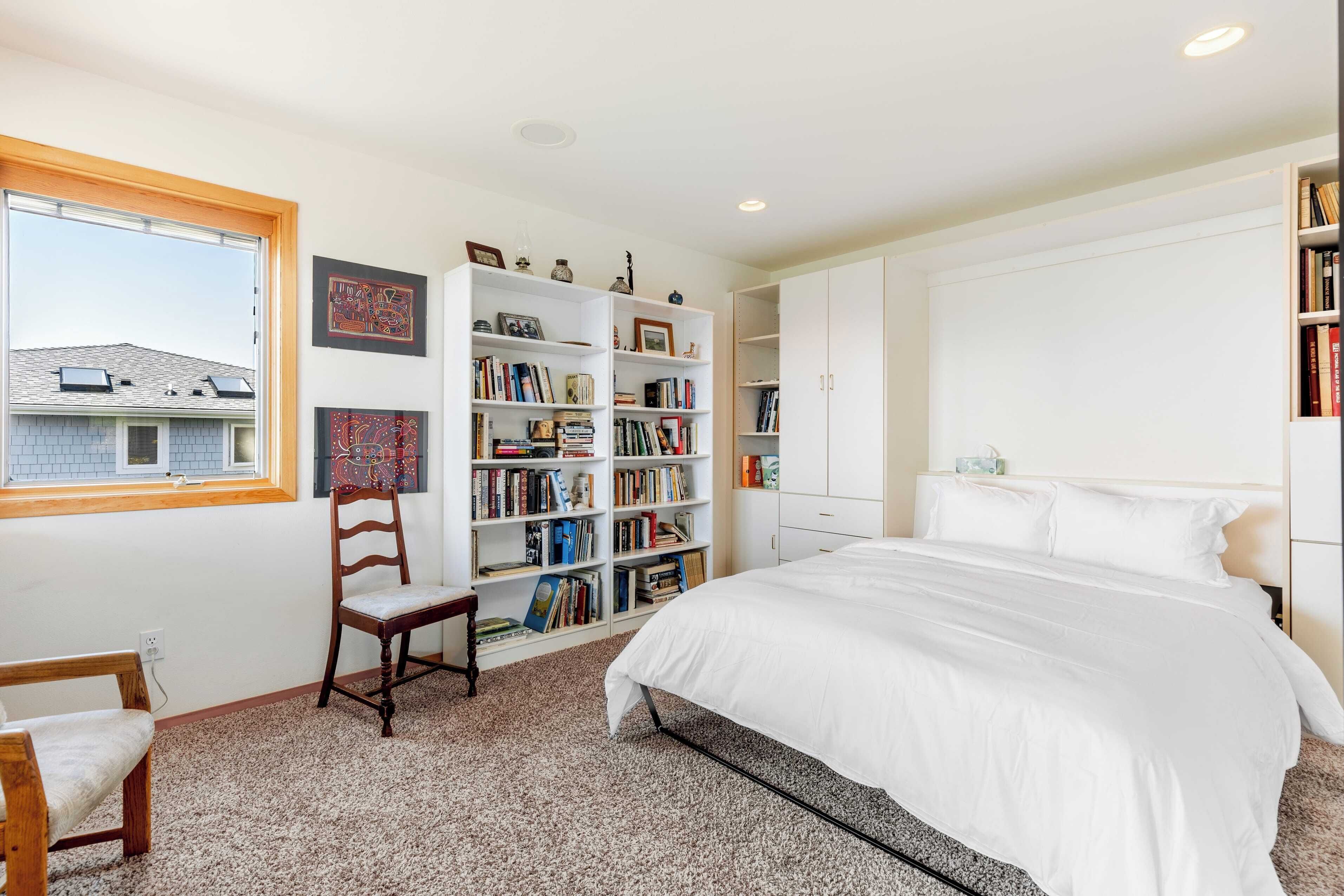 A bedroom with a large bed, a bookshelf, a chair, and a window.