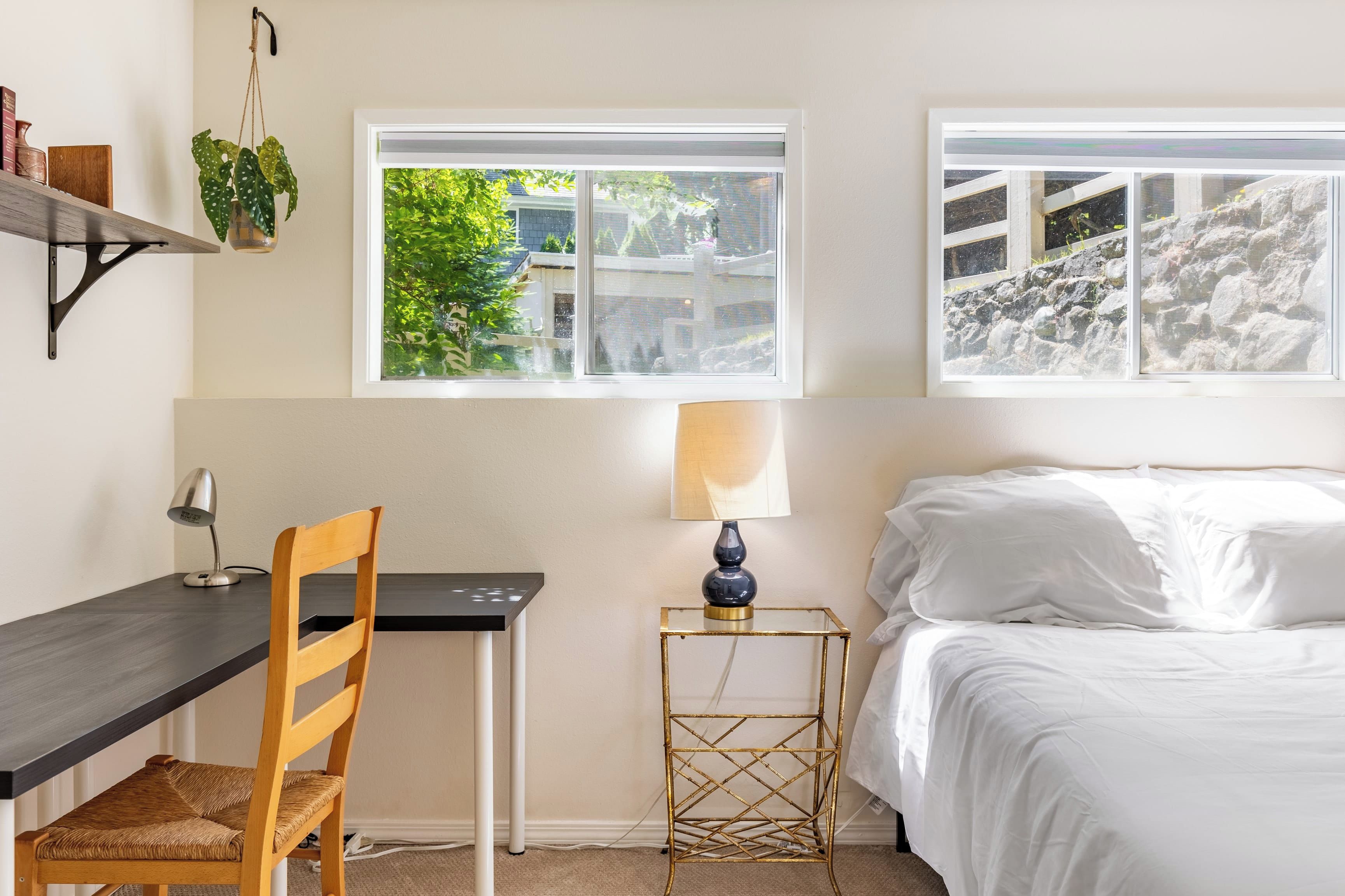 A bedroom with a bed, a desk, a chair, a lamp, and two windows.