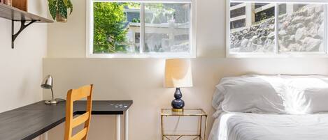 A bedroom with a bed, a desk, a chair, a lamp, and two windows.