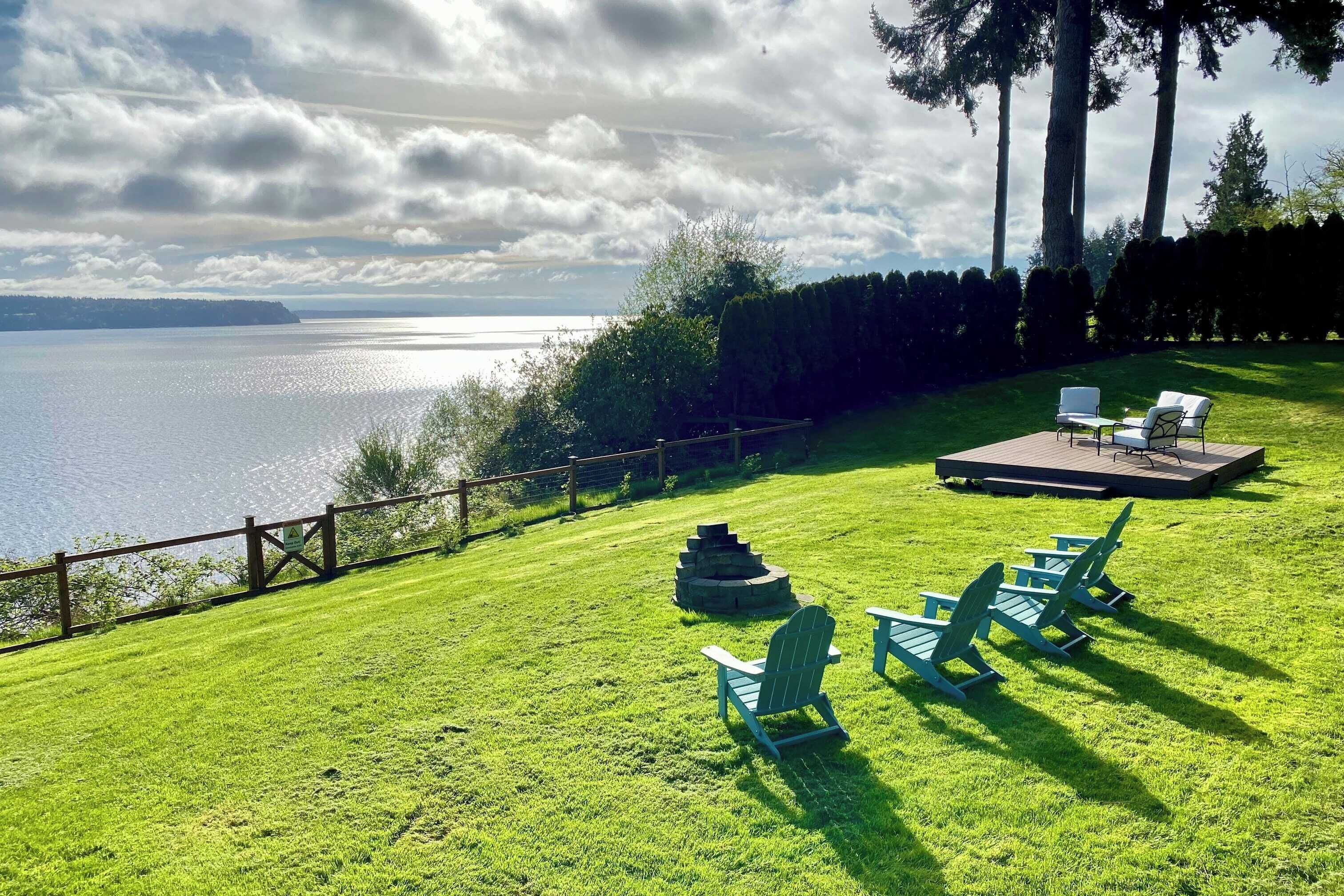 A grassy outdoor area with a view of a body of water, a wooden deck, and Adirondack chairs.