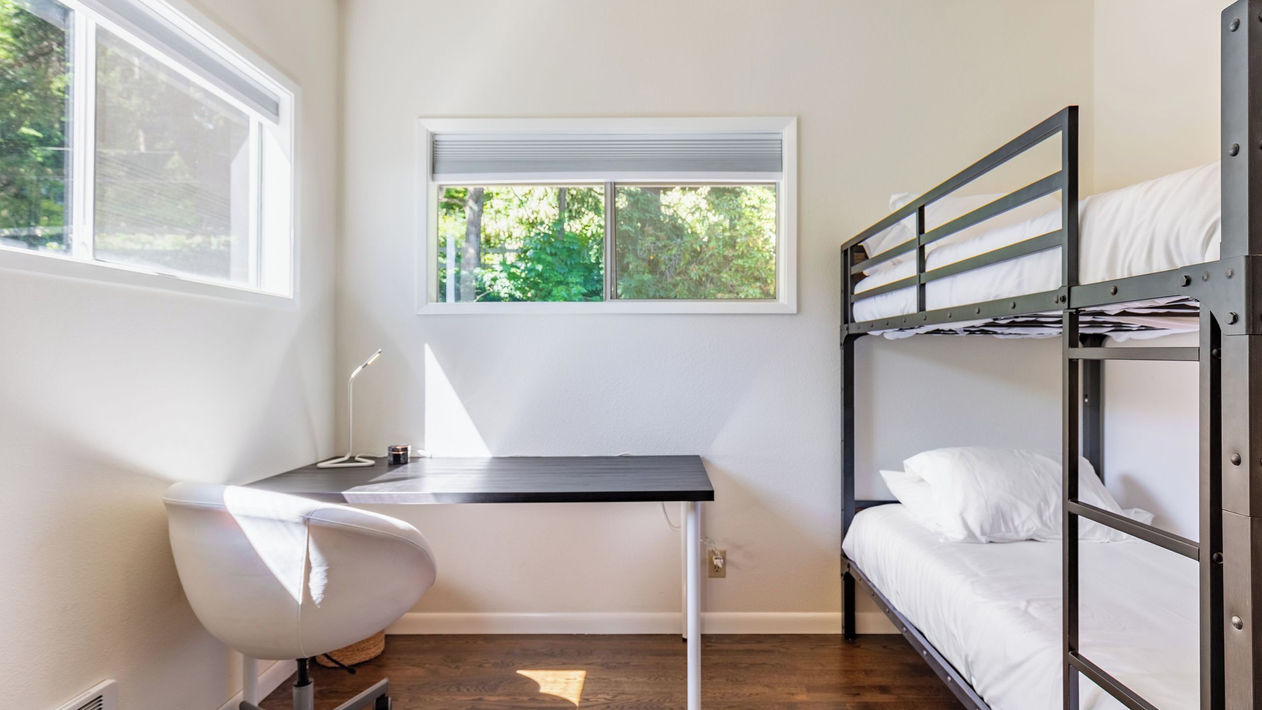 A bunk bed with white bedding, a desk with a chair, and a window with blinds.