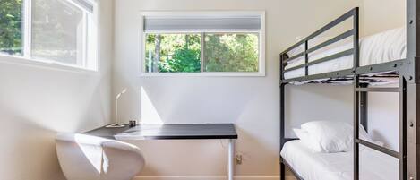 A bunk bed with white bedding, a desk with a chair, and a window with blinds.