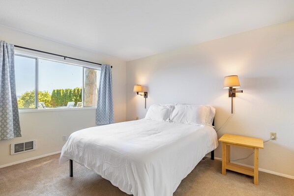 A bedroom with a bed, two wall-mounted lamps, a small wooden nightstand, a window with curtains, and a radiator.