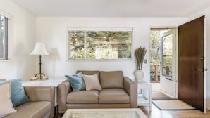A living room with a beige sofa, a small side table, a lamp, and a window with a view of trees. - Eagles Watch by Avantstay Large Deck w/ Views (Coupeville)