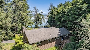 A two-story house with a sloped roof, surrounded by dense forest and a lake view.
