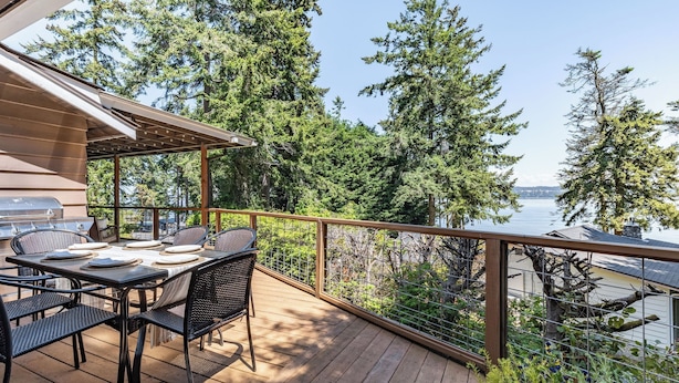 A deck with a grill, dining table, and chairs overlooking a lake with trees. - Eagles Watch by Avantstay Large Deck w/ Views (Coupeville)