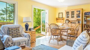 A cozy living room with a comfortable chair, a dining table with chairs, a bookshelf, and a view of greenery outside. - Smugglers Lagoon Beach Cottage by Avantstay (Greenbank)