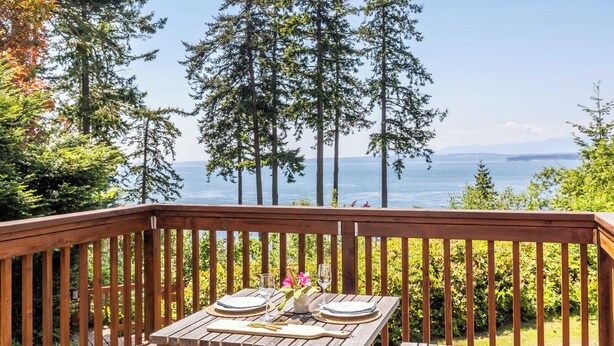 A wooden deck with a table set for two, overlooking a body of water and tall trees. - Smugglers Lagoon Beach Cottage by Avantstay (Greenbank)