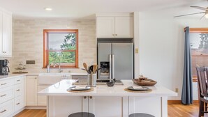 Une cuisine moderne avec des meubles blancs, un îlot central intégré, des appareils en acier inoxydable et une fenêtre donnant sur la verdure.