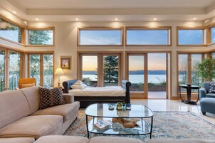 A spacious living room with a large bed, a sofa, a coffee table, and a view of a lake through large windows.