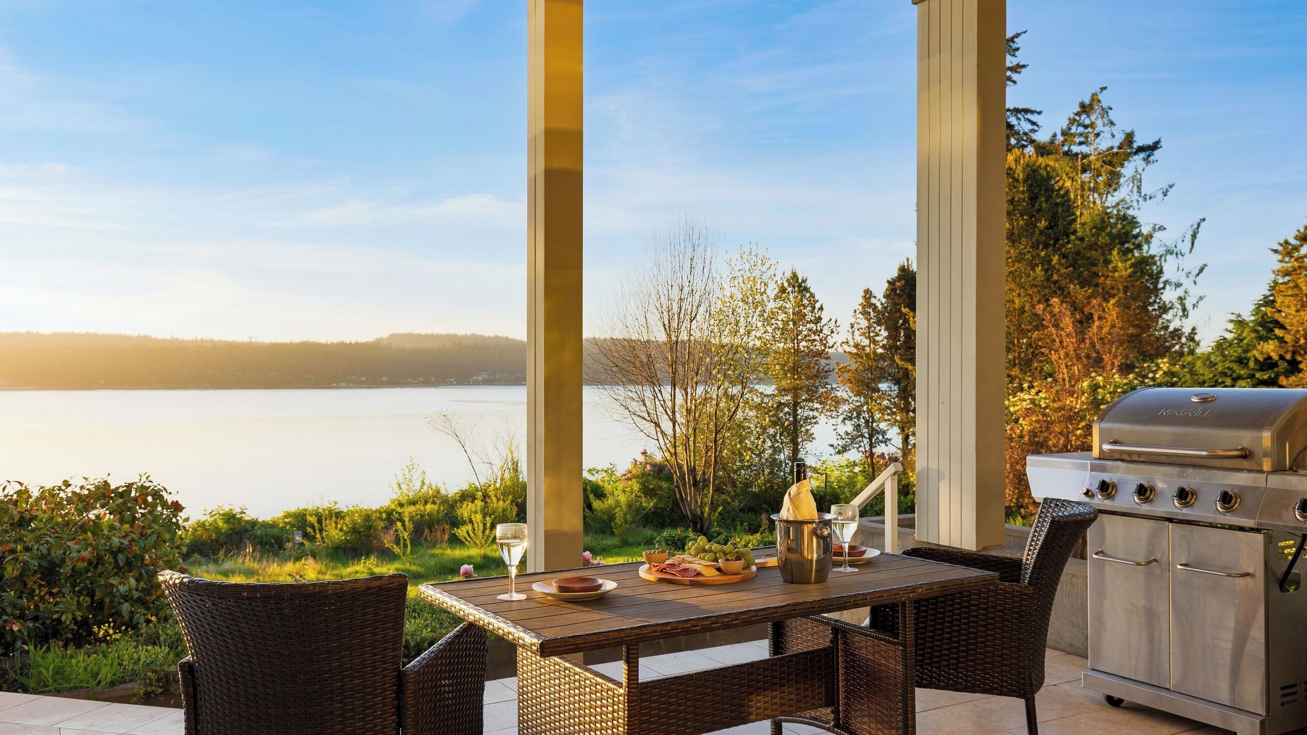 An outdoor dining area with wicker chairs, a table set with plates, a grill, and a view of a lake.