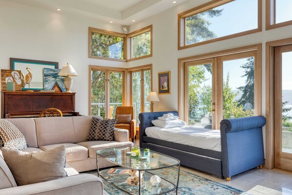 A spacious living room with a sofa, a glass coffee table, and a bed with a blue headboard. Large windows allow natural light and views of greenery. - Orca Ridge on Resort Road by Avantstay A+ Views (Greenbank)