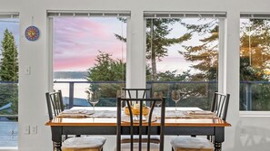 A dining table with chairs, a bowl, wine glasses, and a view of a lake and trees through large windows. - Incredible Water Views, Deck Relaxing Retreat Greenbank Getaway by Avantstay (Greenbank)