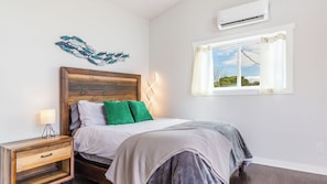 A bedroom with a wooden headboard, a bedside table with a lamp, a window with curtains, and a wall-mounted air conditioner. - Chateau Coho by Avantstay Easy Beach Access (Greenbank)