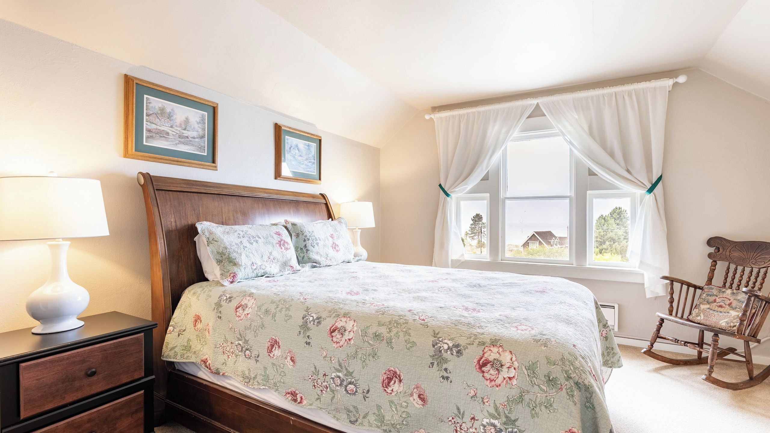A bedroom with a wooden bed, two bedside lamps, a rocking chair, and a window with curtains.