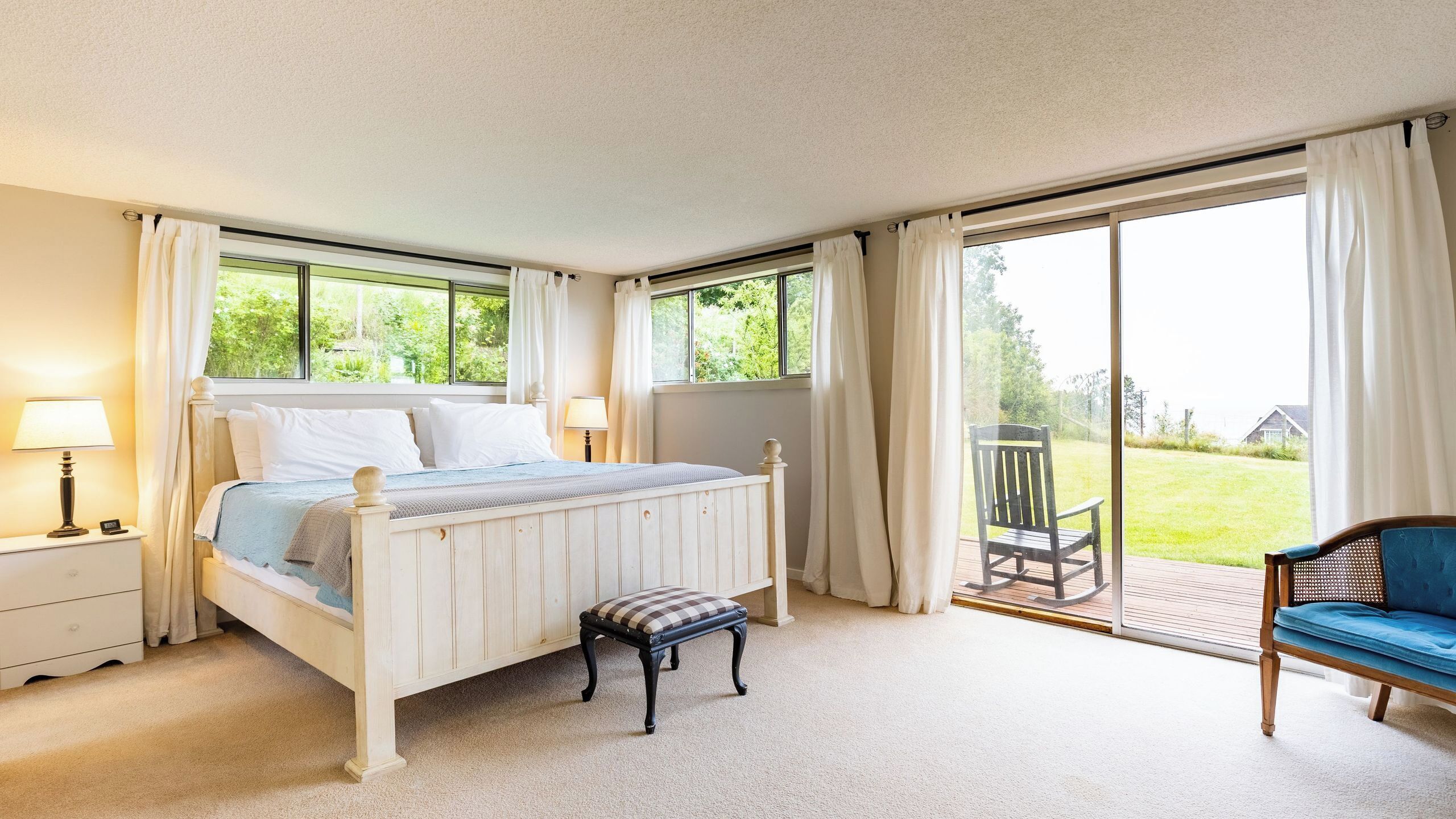 A bedroom with a bed, bedside lamps, a bench, a chair, and a view of a garden through a sliding door.