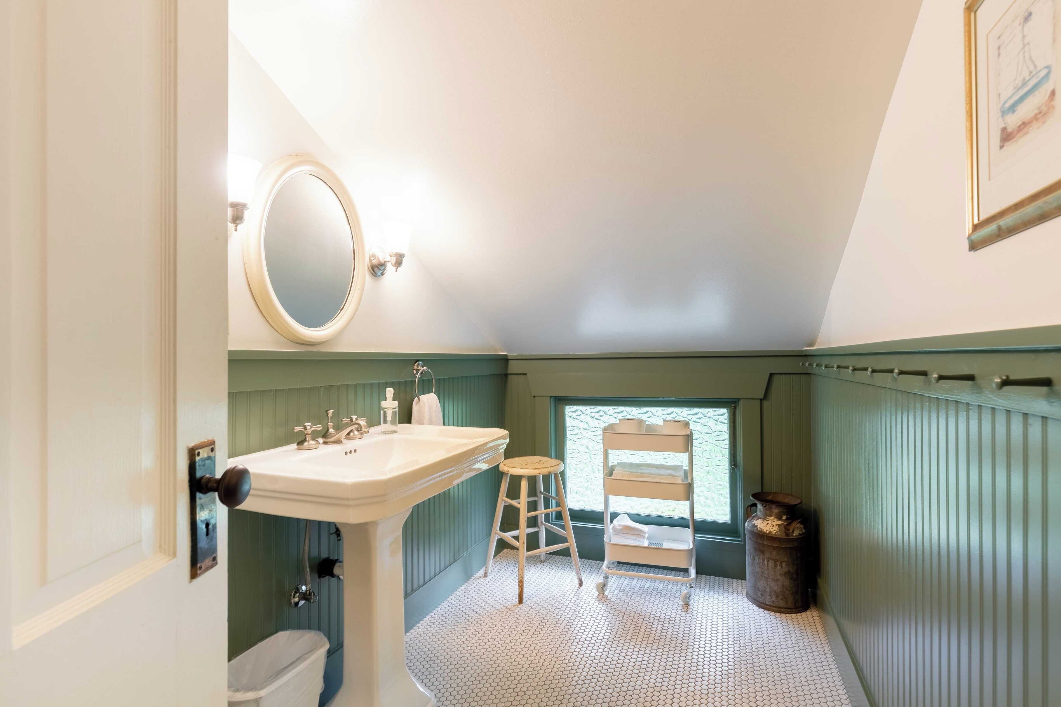 A bathroom with a pedestal sink, a mirror, a stool, a shelf with towels, and a window.