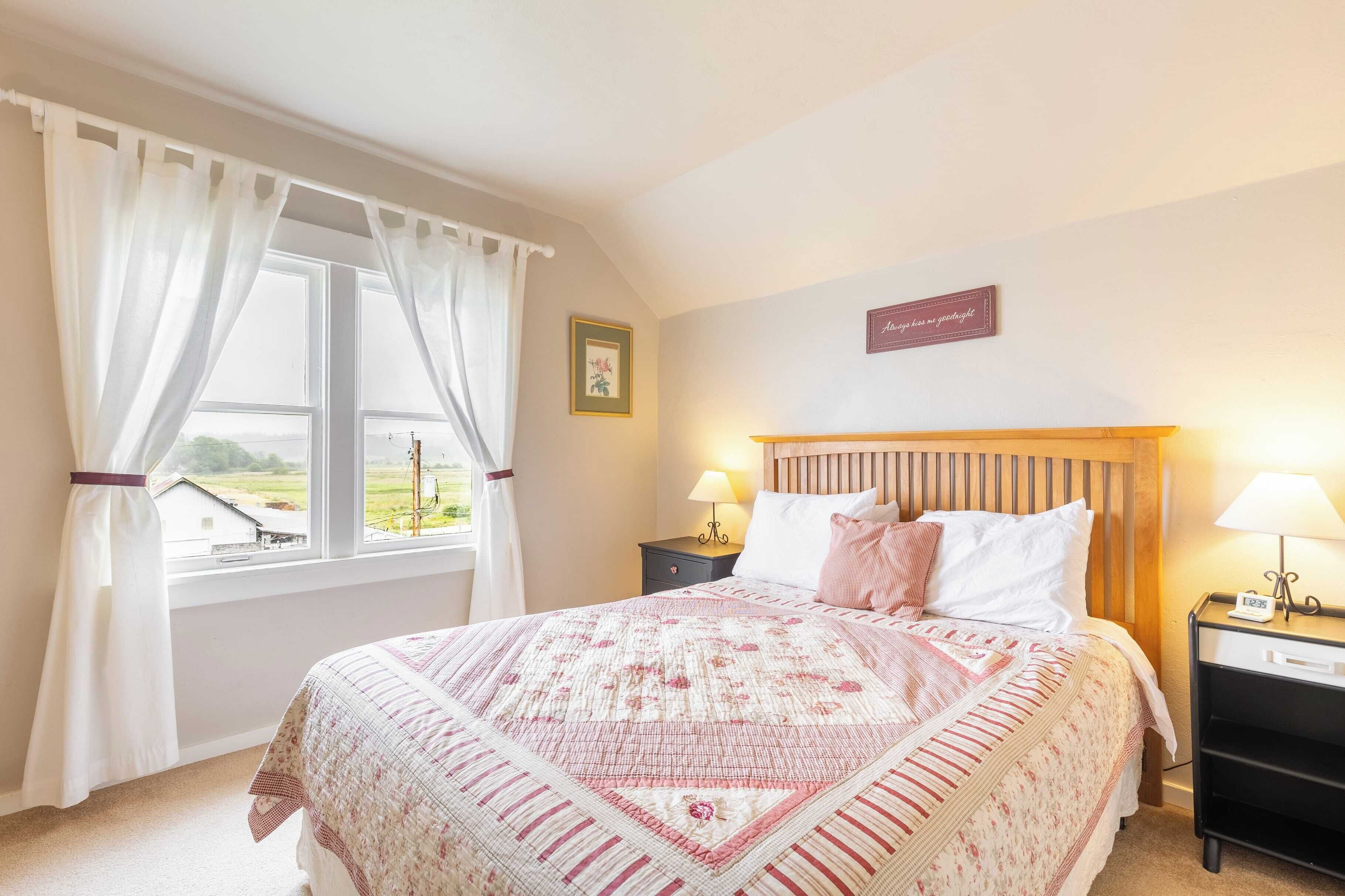 A bedroom with a bed, two bedside tables, a nightstand, a window with curtains, and a wall-mounted sign.