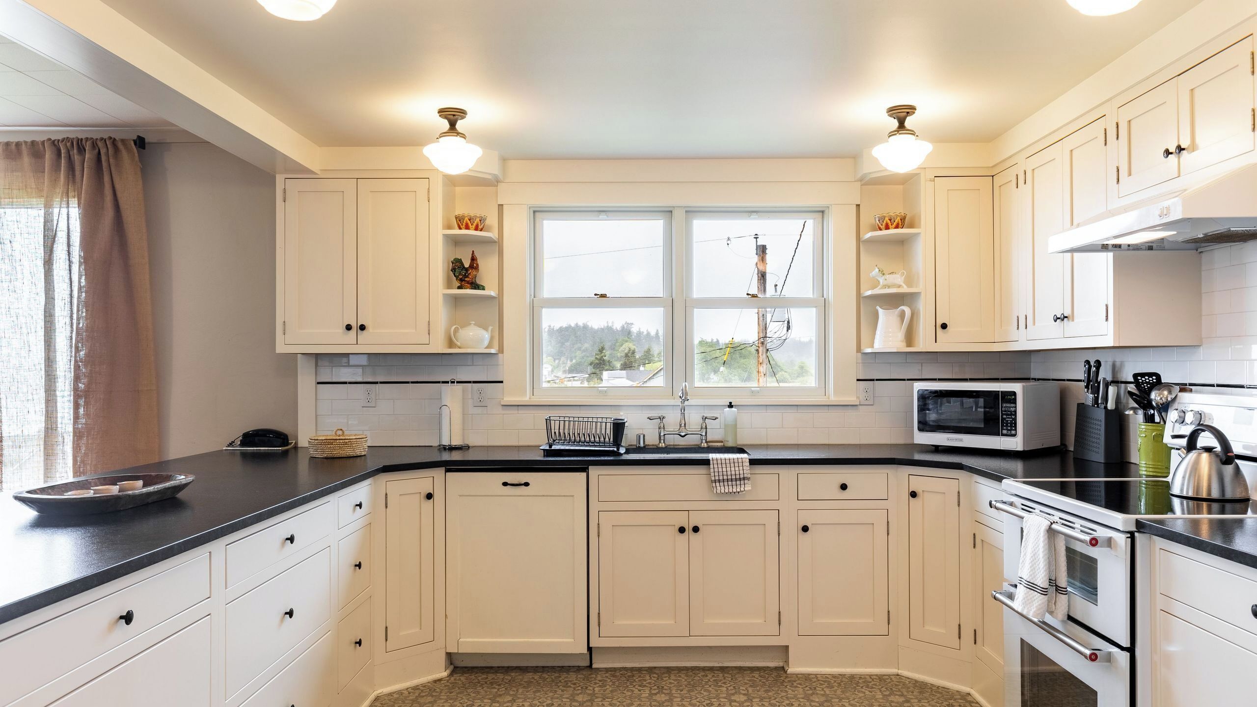 Una cocina con armarios blancos, microondas, horno y ventana con vistas a árboles.