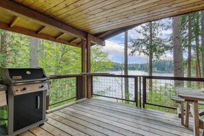 Eine Holzterrasse mit Grill und Tisch, mit Blick auf einen von Bäumen umgebenen See.