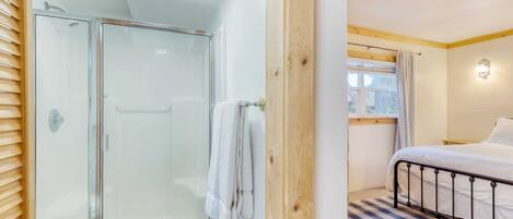 A bedroom with a bed, a window with curtains, a wooden door, and a shower area with a glass door.