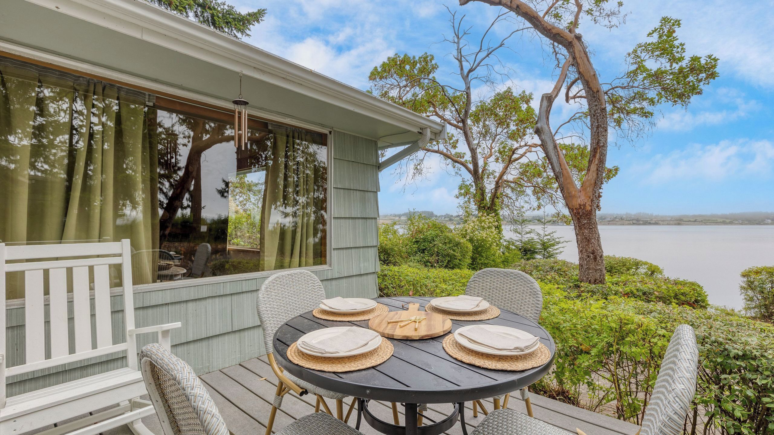 A deck with a round table set for four, surrounded by wicker chairs, overlooking a body of water.