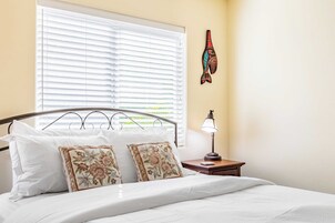 A neatly made bed with white linens and floral pillows, a bedside lamp, and a window with blinds.