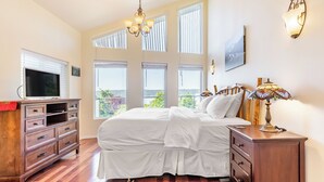 Un dormitorio con cama, cómoda, televisor y ventanas amplias.