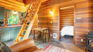 Interior acolhedor de uma cabana, com decoração em madeira, uma sala de jantar com mesa e cadeiras, uma cama e uma lareira.