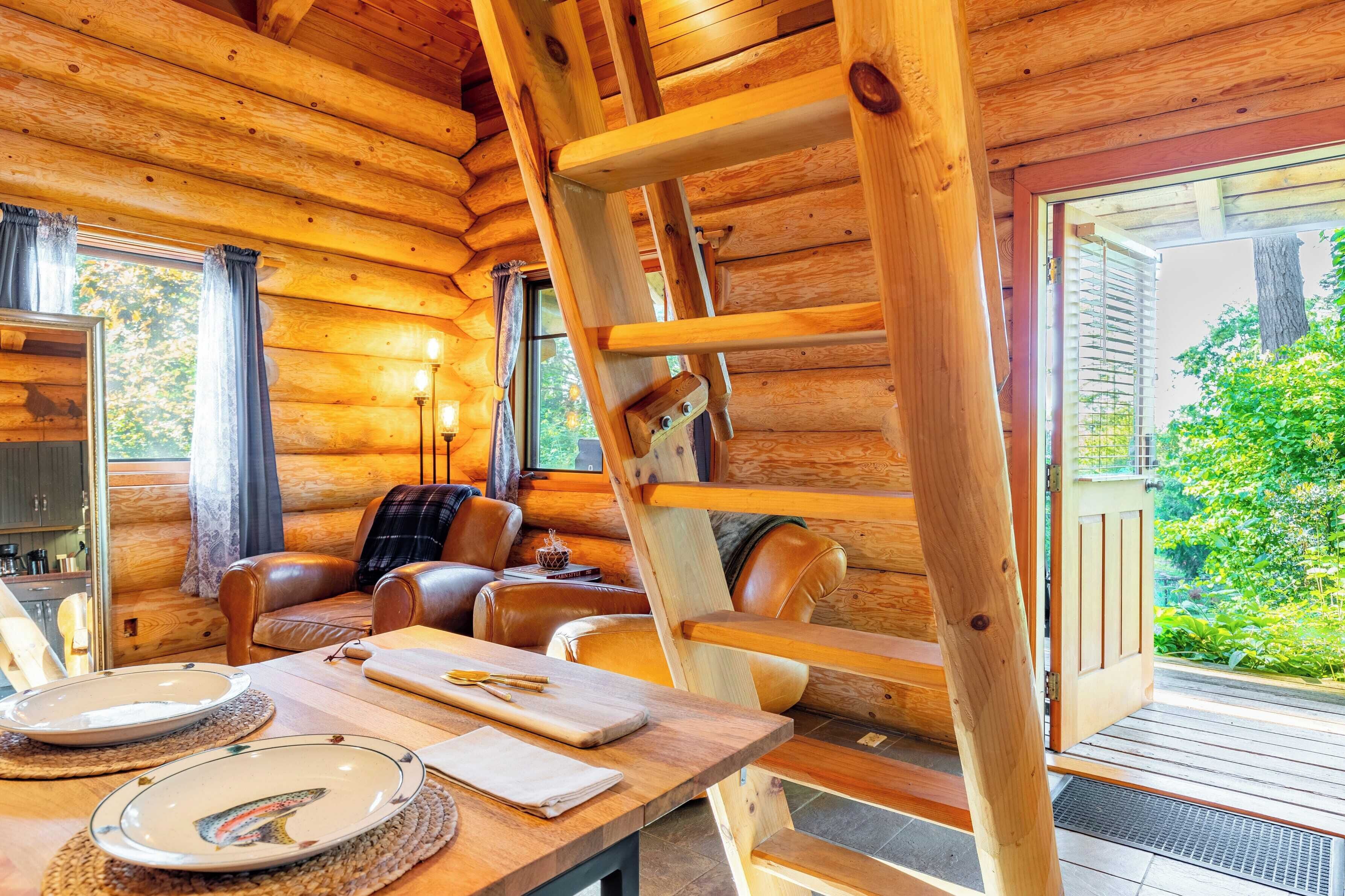 A log cabin interior with a dining table set, a ladder, leather sofas, and a view of greenery outside.