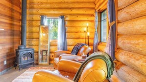 Interior de uma cabana de tronco com um sofá de couro, uma lareira e vista para o exterior.