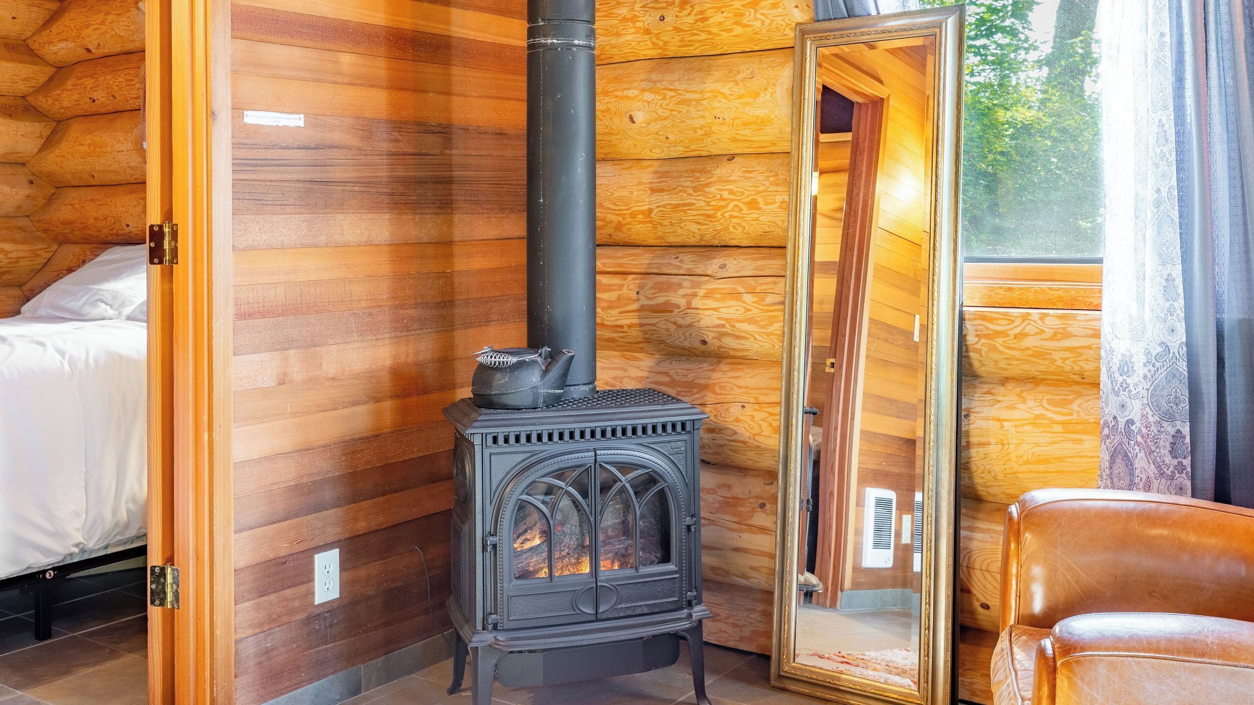 A log cabin room with a wood-burning stove, a bed, and a leather chair.