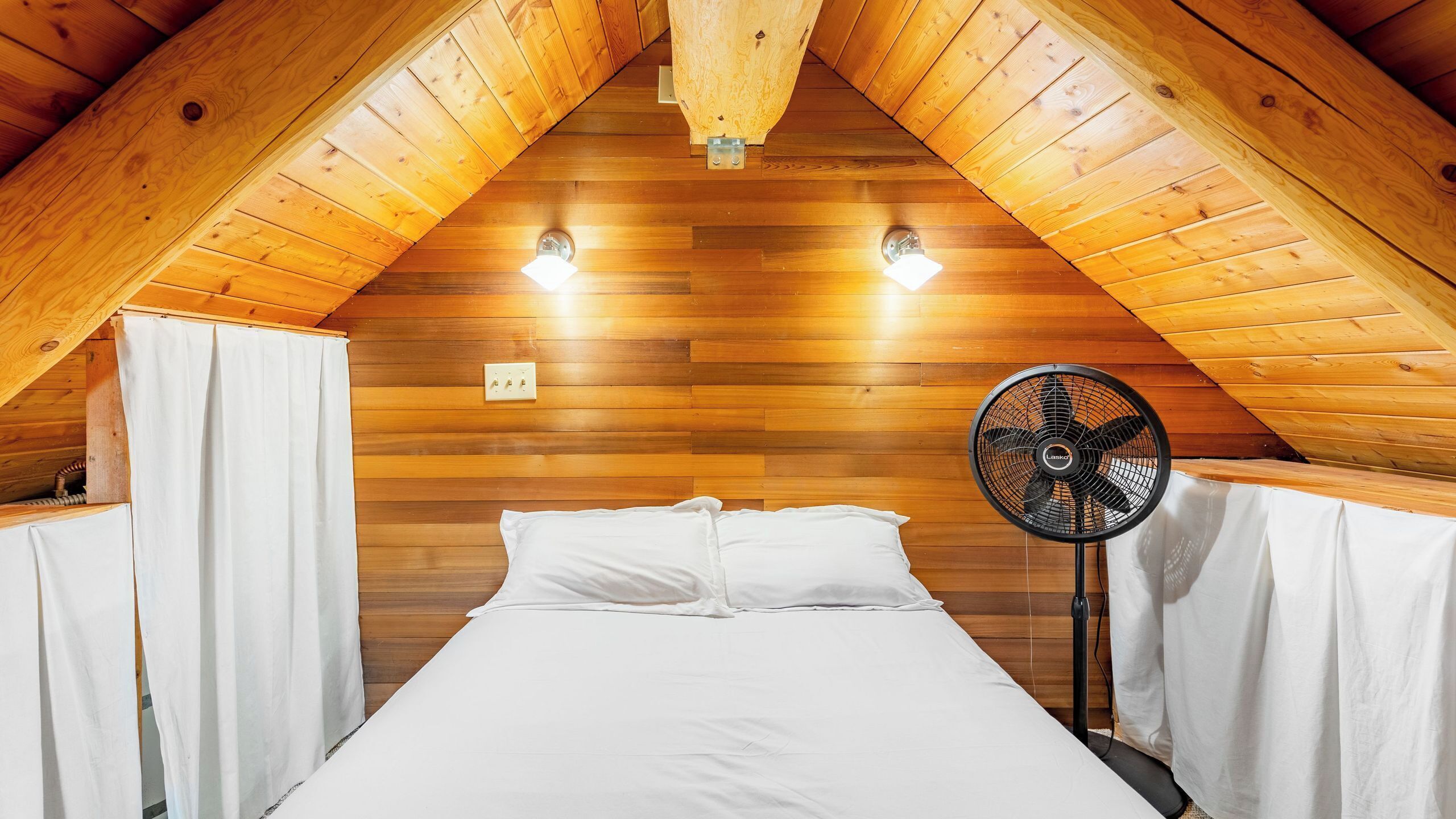 A bedroom with a sloped ceiling, wooden walls, a bed with white bedding, a standing fan, and two wall-mounted lights.