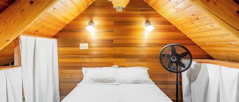 A bedroom with a sloped ceiling, wooden walls, a bed with white bedding, a standing fan, and two wall-mounted lights.