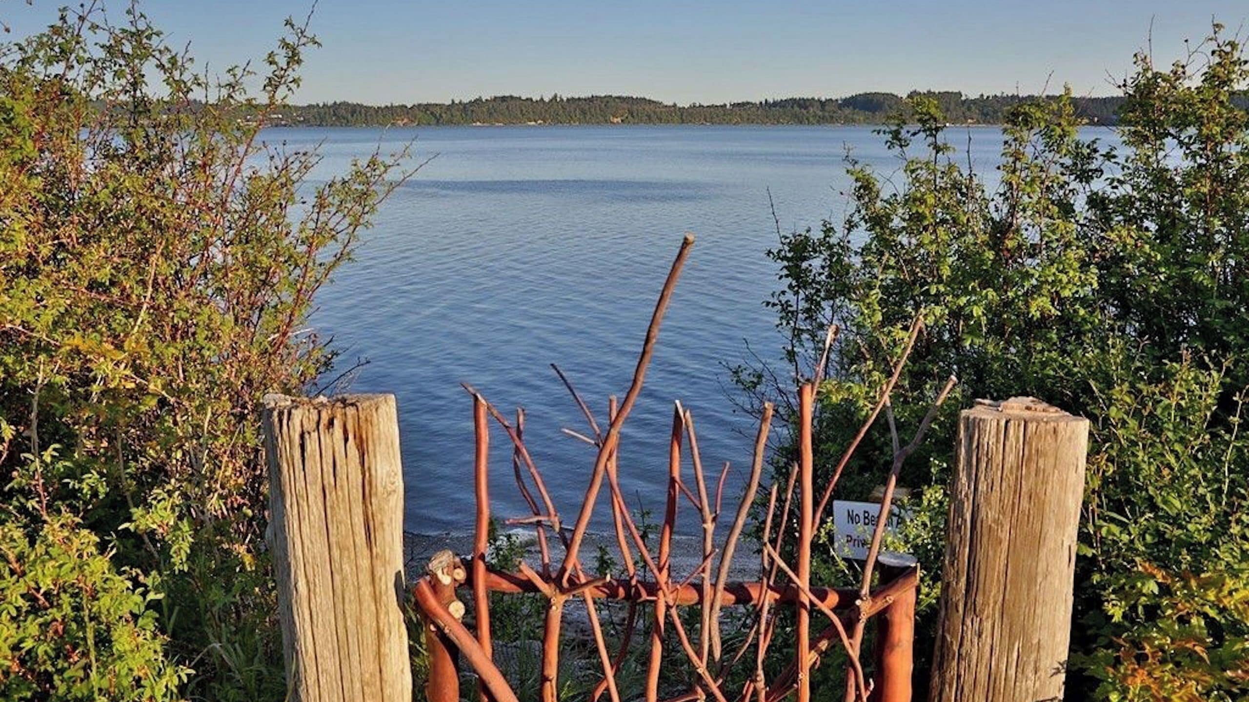 Une clôture en bois avec un panneau, un lac paisible et une rive bordée d’arbres.