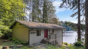 A small cabin with a red door, a porch, and a view of a lake surrounded by trees.