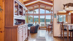 A wooden interior with a dining area, kitchen, and large windows overlooking a forested area.
