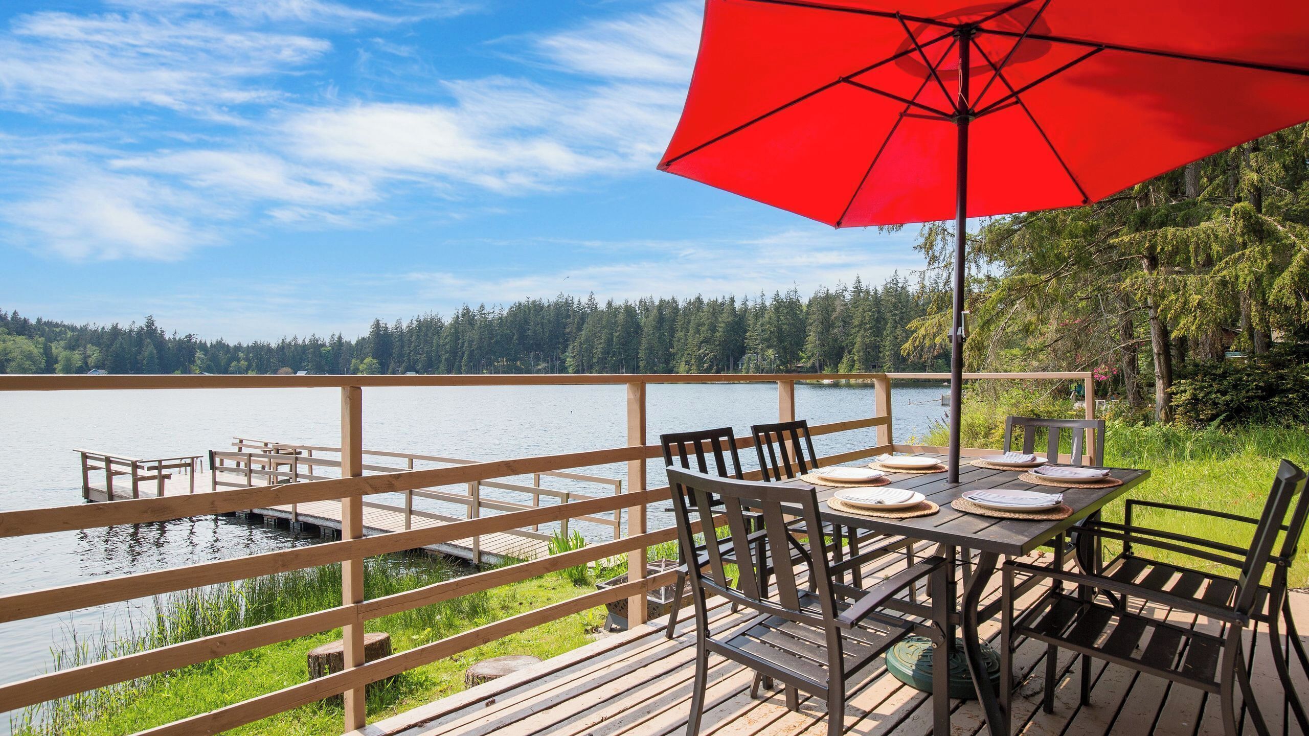 Una terraza de madera con una mesa y sillas, con vistas a un lago bordeado por un bosque.