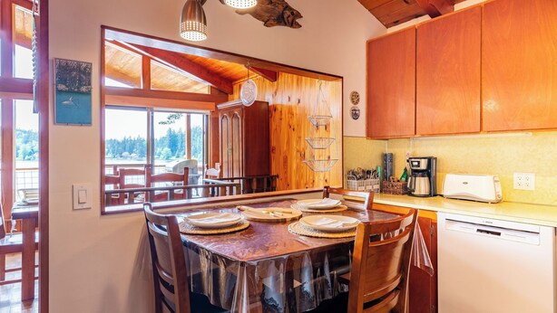 A wooden interior with a dining table set for four, a kitchen with a coffee maker, and a view of the outdoors. - Goss Lake Getaway by Avantstay Lakefront Escape (Langley)