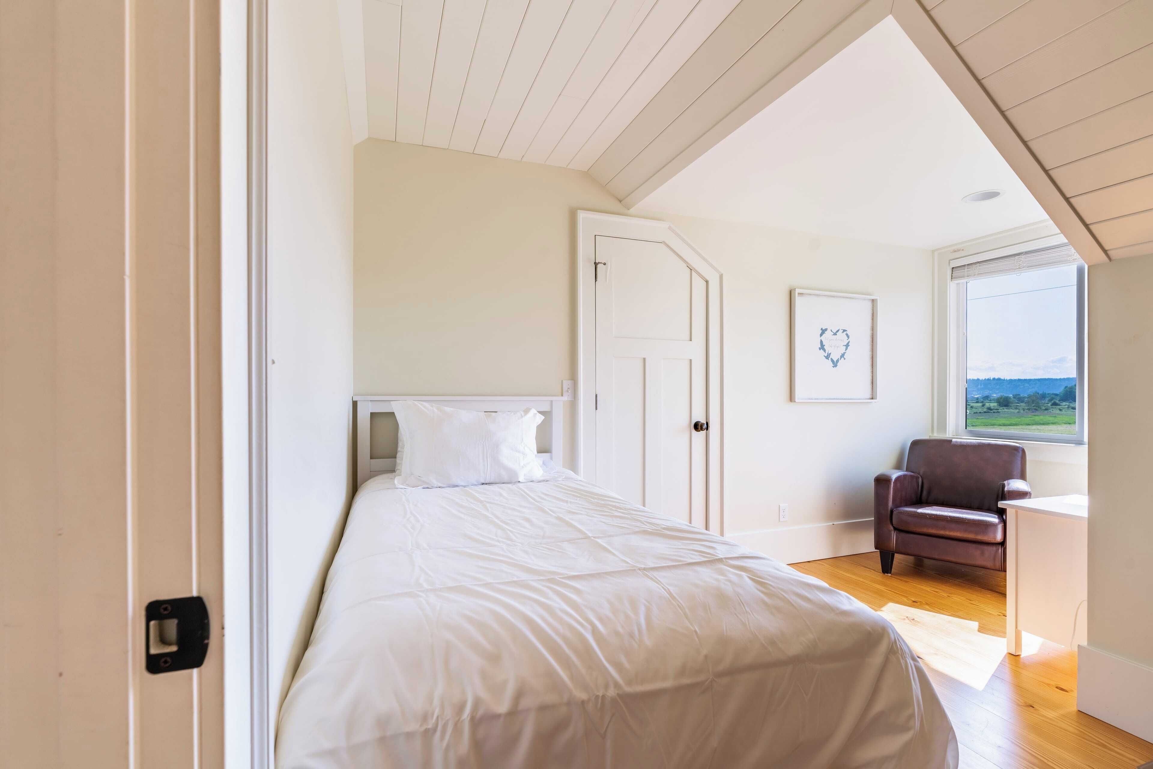 A bedroom with a bed, a chair, a desk, a window with a view, and a door.