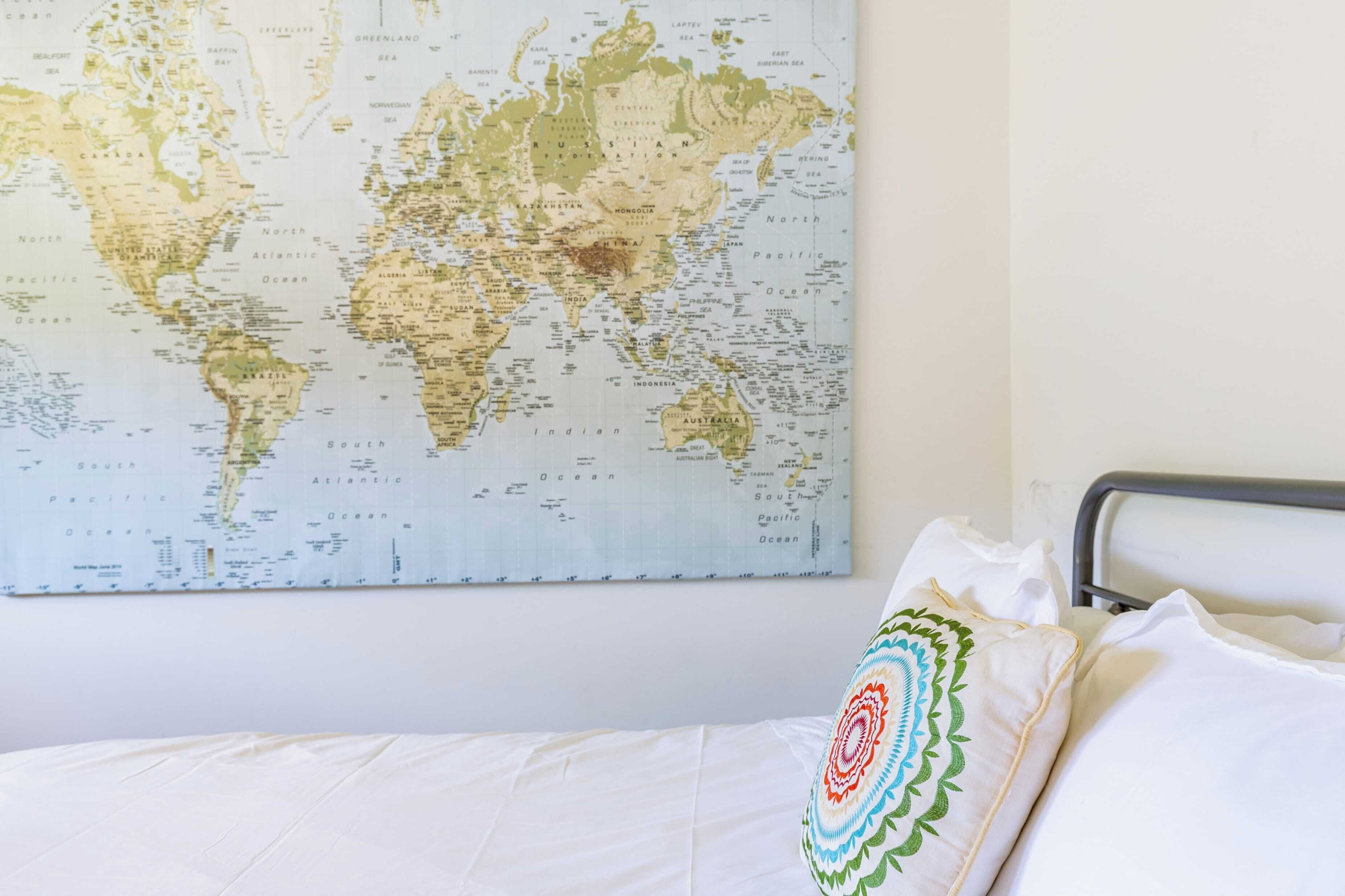 Cama com roupa de cama branca e um travesseiro decorativo, um mapa-múndi na parede e cabeceira de metal.