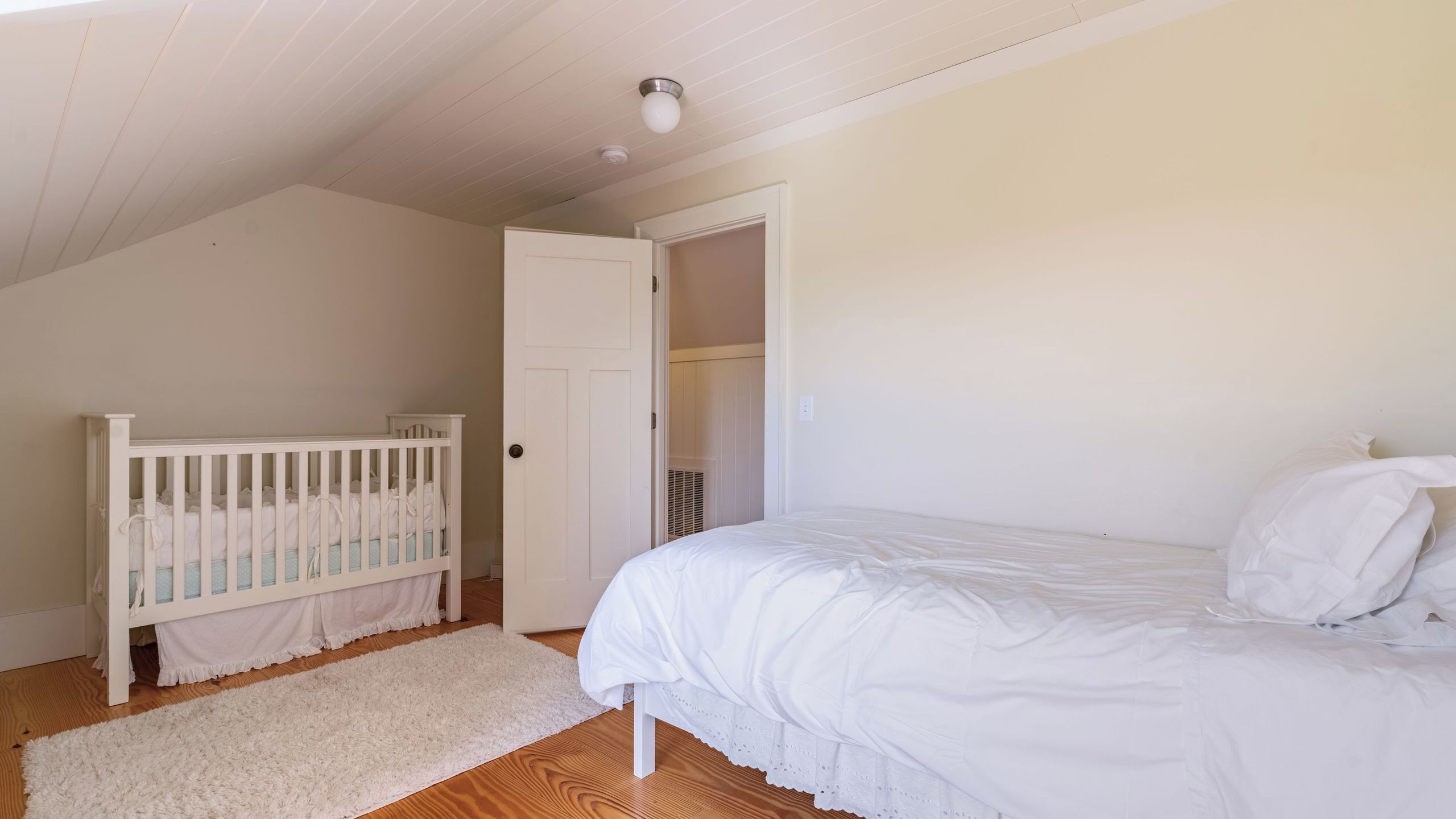 Ein Schlafzimmer mit einem Kinderbett, einem Bett, einer weißen Tür und einer Deckenleuchte.